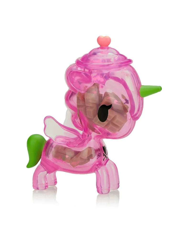 The tokidoki Sugar Shop Unicorno Blind Box features a pink translucent collectible with a green horn and tail, white wings, a heart on its head, and is filled with small brown blocks.