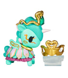 A tokidoki Unicorno After Dark Masquerade Blind Box figurine with teal, gold, and purple accents stands beside a gold crown with a snake motif, both displayed as collectible toys on a clear stand.