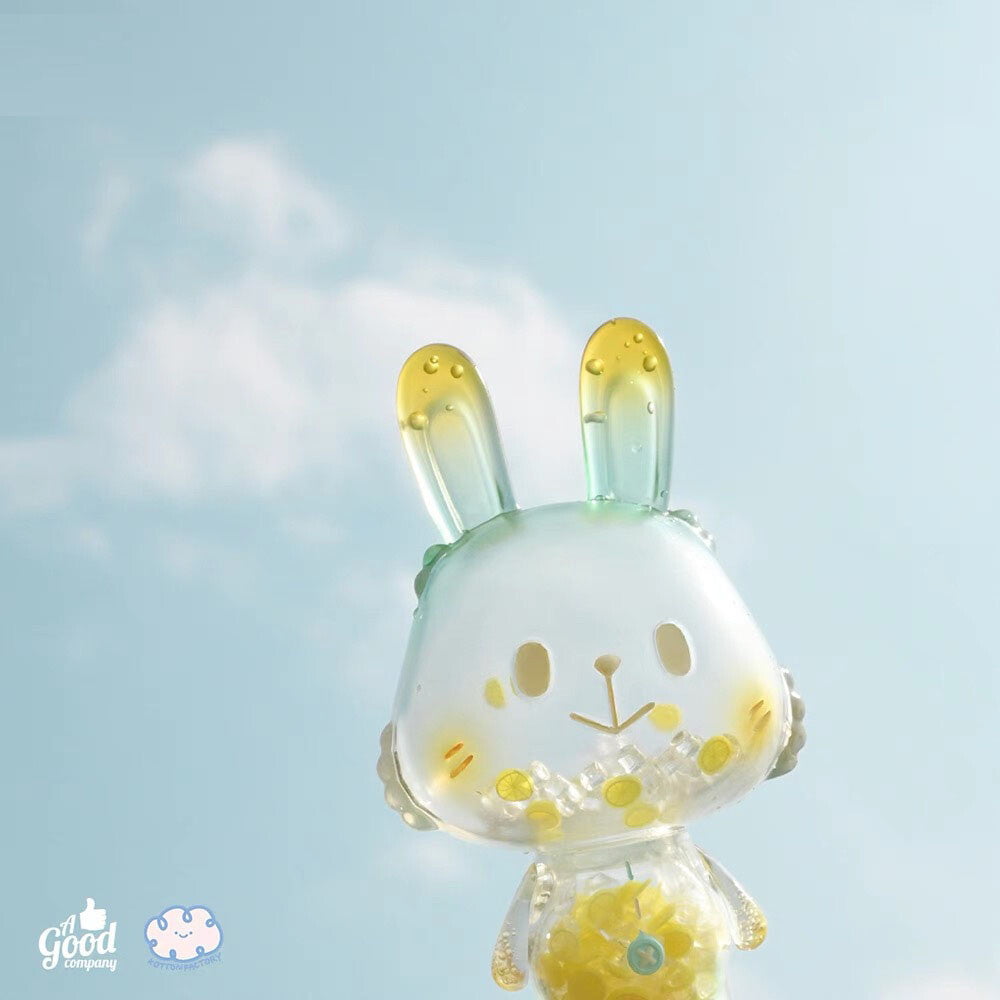 Lemon Soda Robin by A Good Company—a translucent bunny figure with yellow and green accents—stands out against a blue sky with scattered clouds, making it a perfect shelf ornament for charming home decor. Logos are in the lower-left corner.
