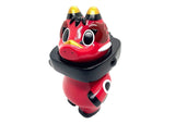 The Teresa Chiba Akabegon- Red is a red and black cartoon-style figurine inspired by Japanese Akabeko folklore, featuring large eyes, yellow-tipped horns, a black collar, and stands upright on a white background.