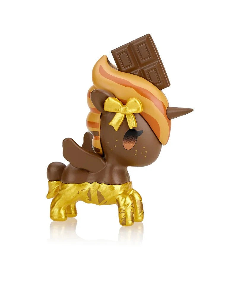 The tokidoki Toki Mochi Donut Unicorno Blind Bag features a squishy Unicorno toy with a chocolate brown body, gold legs, caramel-striped mane, gold bow, and chocolate bar atop its head against a white background.