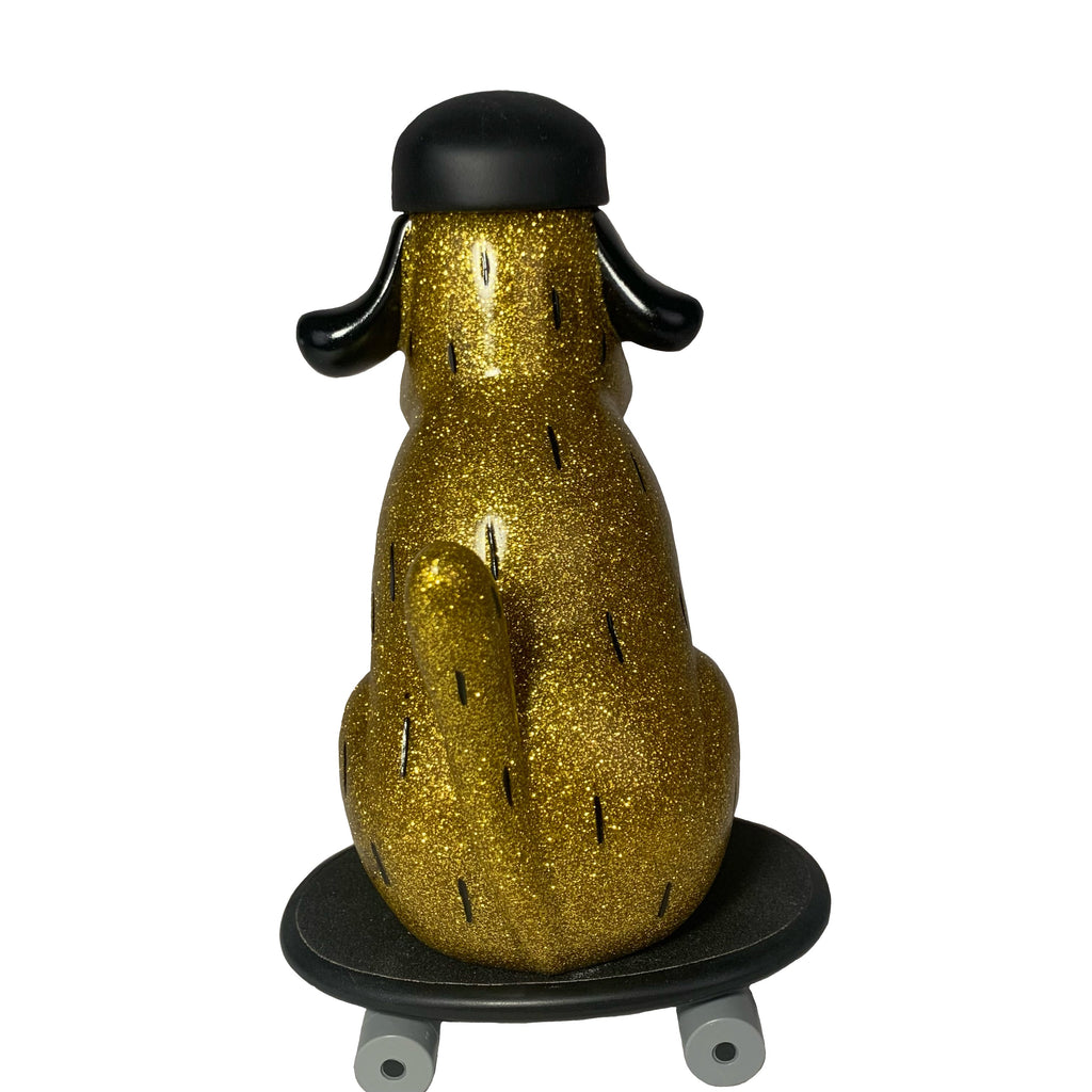 The How2Work One Chu Sk8- Gold figure features gold glitter, black ears, and a helmet as it sits on a black skateboard, seen from behind.