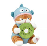 A Kitan Club Mofusand x Sanrio Series 2 Capsule figurine features an orange tabby cat in a blue costume with ears, holding a kiwi slice—perfect for Mofusand kitty fans.