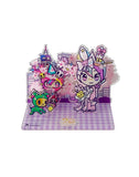 The tokidoki 20th Anniversary Acrylic Standee Blind Box features vibrant cartoon characters with party hats and pastel accents on a purple checkered base, set against a cityscape and cherry blossoms, celebrating 20 years of tokidoki.