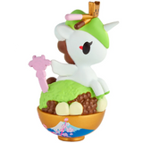A cute tokidoki Shaved Ice Dreams Unicorno Blind Box collectible features a unicorn figure with a green hat sitting atop a dessert bowl adorned with green topping, round brown balls, and decorative sticks. The bowl showcases an adorable cherry blossom and mountain design, inspired by the Shaved Ice Dreams Unicorno series.
