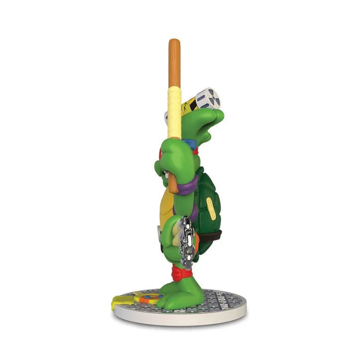 A side view of the Kidrobot Teenage Mutant Ninja Turtles TMNT GURU Resin Figure by CHOGRIN, holding a staff and standing on a round, colorful base—an eye-catching collectible for TMNT fans.