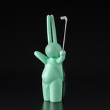 The Daily Flasher by mr. clement from Tomenosuke Shoten is a mint green, glow-in-the-dark vinyl toy rabbit holding a selfie stick, shown facing away against a black background.
