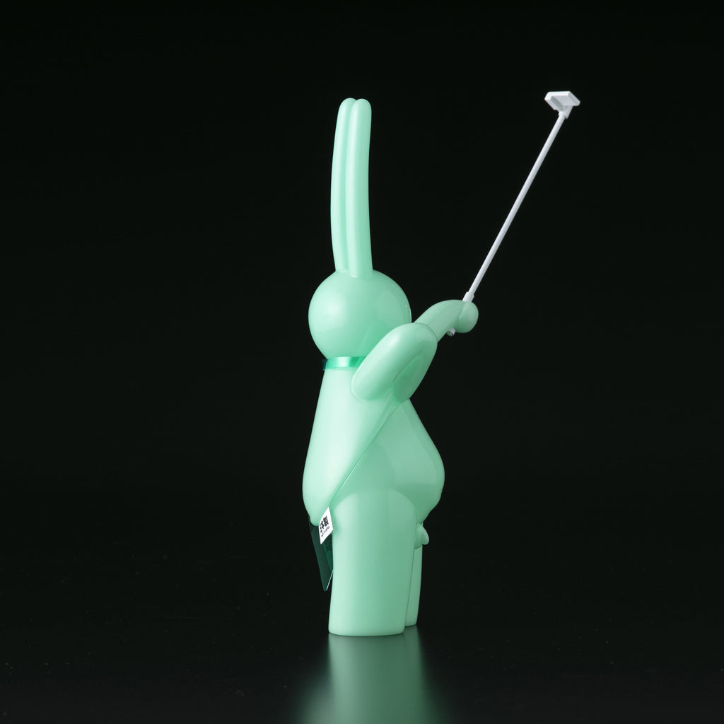 The Daily Flasher by mr. clement—a mint-green, glow-in-the-dark vinyl rabbit toy from Tomenosuke Shoten—stands upright holding a selfie stick, making it a playful collectible against a black background.