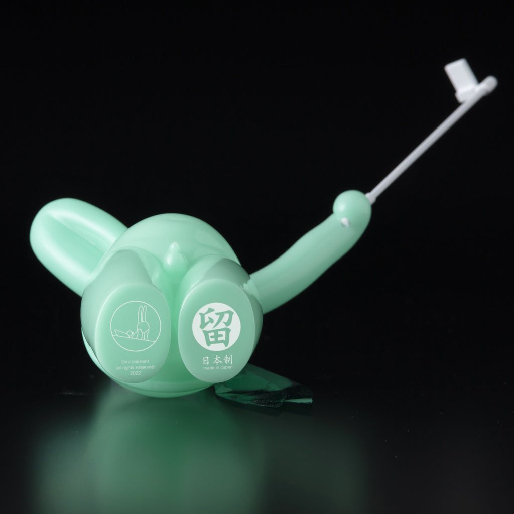 The Daily Flasher by mr. clement for Tomenosuke Shoten is a mint green plastic piece with a round base, long handle, and thin white rod, resembling a glow-in-the-dark vinyl toy, set against a black background.