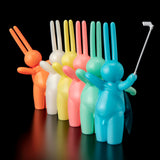 Seven vibrant The Daily Flasher figurines by mr. clement stand in a row, the front one with a selfie stick. These playful collectibles from Tomenosuke Shoten are arranged against a bold black background.