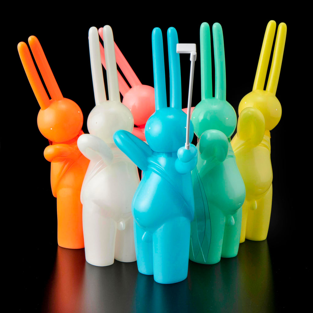 Six vibrant, bunny-shaped The Daily Flasher figures by mr. clement for Tomenosuke Shoten—including a glow-in-the-dark vinyl—stand together on a black background, with the blue one in front holding a selfie stick.