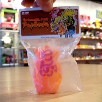The Popsoda Finger Puppet - Orange by Pop Soda is shown in a clear plastic bag on a wooden surface, with colorful shelves in the background.