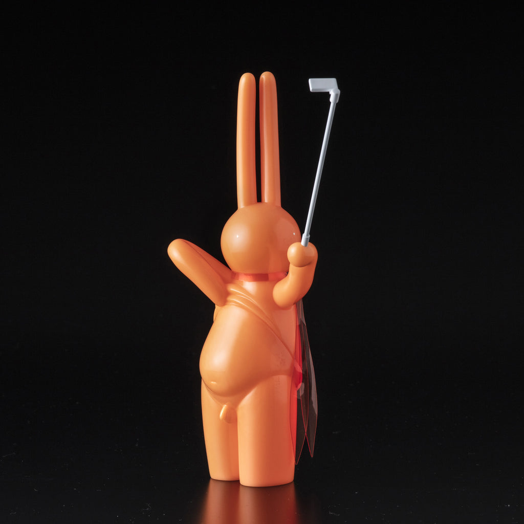 The Daily Flasher by mr. clement for Tomenosuke Shoten features an orange sofubi rabbit figurine with a raised selfie stick and a red cape, playfully referencing the brand’s iconic glow-in-the-dark vinyl art scene.