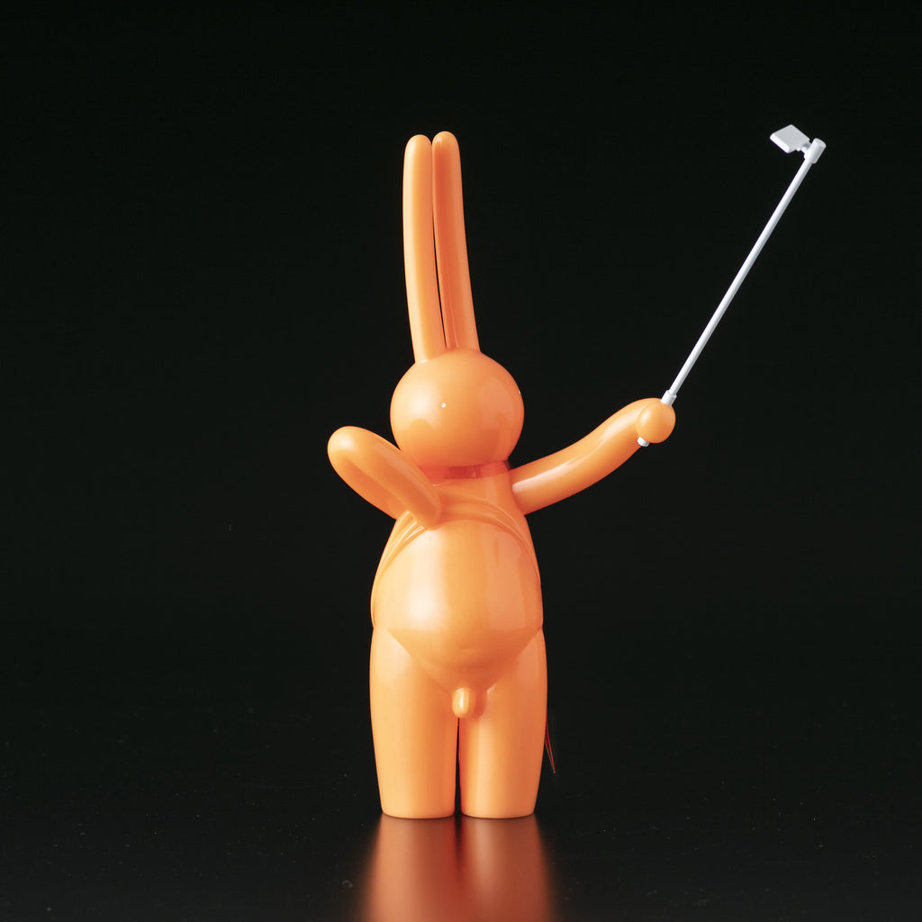 The Daily Flasher by mr. clement for Tomenosuke Shoten stands upright, holding a white selfie stick—an orange, glow-in-the-dark vinyl rabbit figure playfully posed against a black background.
