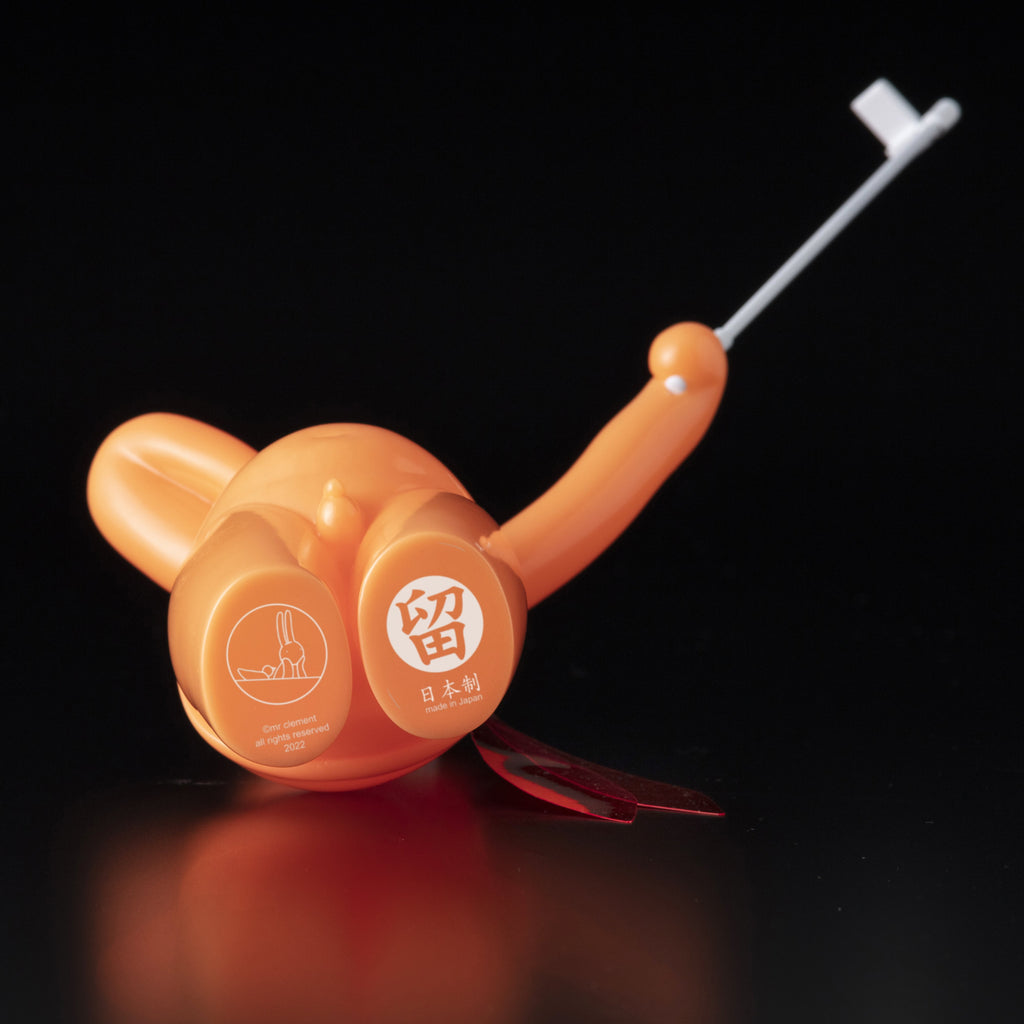 The Daily Flasher by mr. clement, from Tomenosuke Shoten, is an orange plastic device with a retractable metal hook and bilingual labeling in Japanese and English, shown on a black surface—resembling a sofubi collectible.