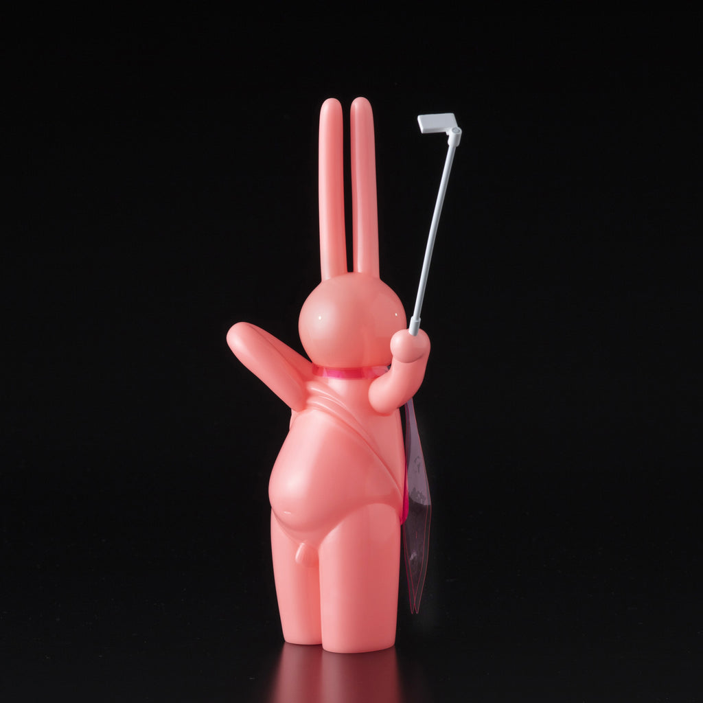 The Daily Flasher by mr. clement, a pink glow-in-the-dark vinyl rabbit from Tomenosuke Shoten, stands against a black background, holding a selfie stick and waving with its left hand.