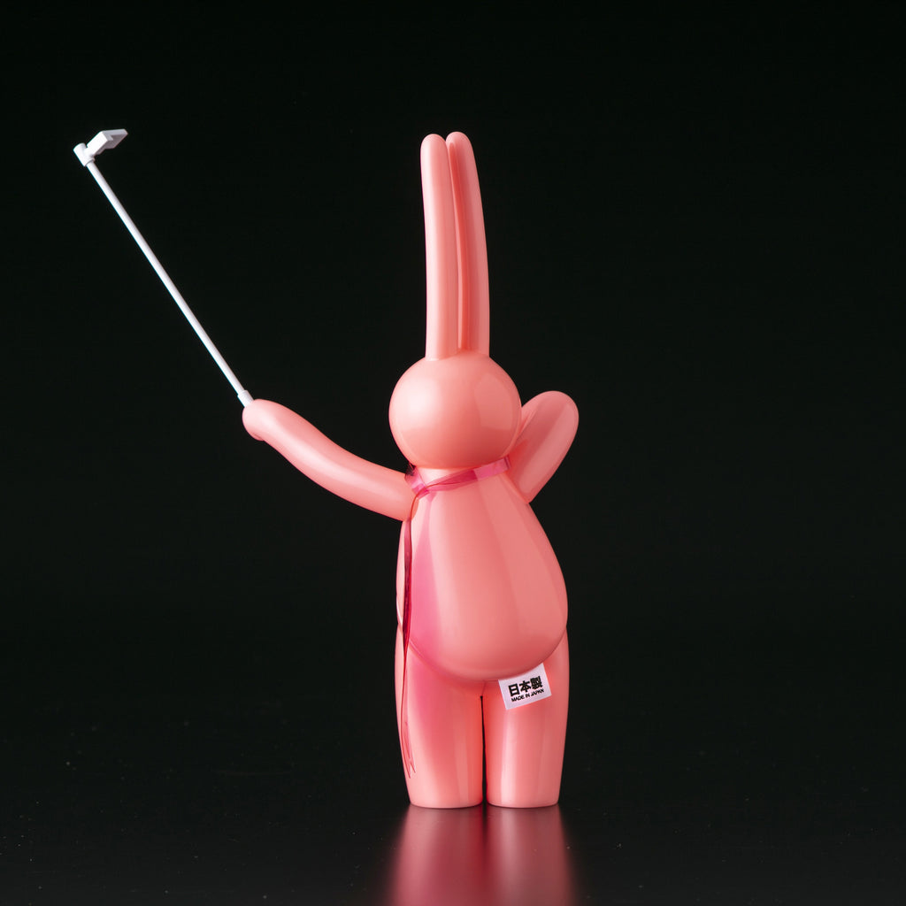 The Daily Flasher by mr. clement is a pink, rabbit-like glow-in-the-dark vinyl figure from Tomenosuke Shoten, shown upright on a black background holding a selfie stick in its right paw—a standout collectible for enthusiasts.
