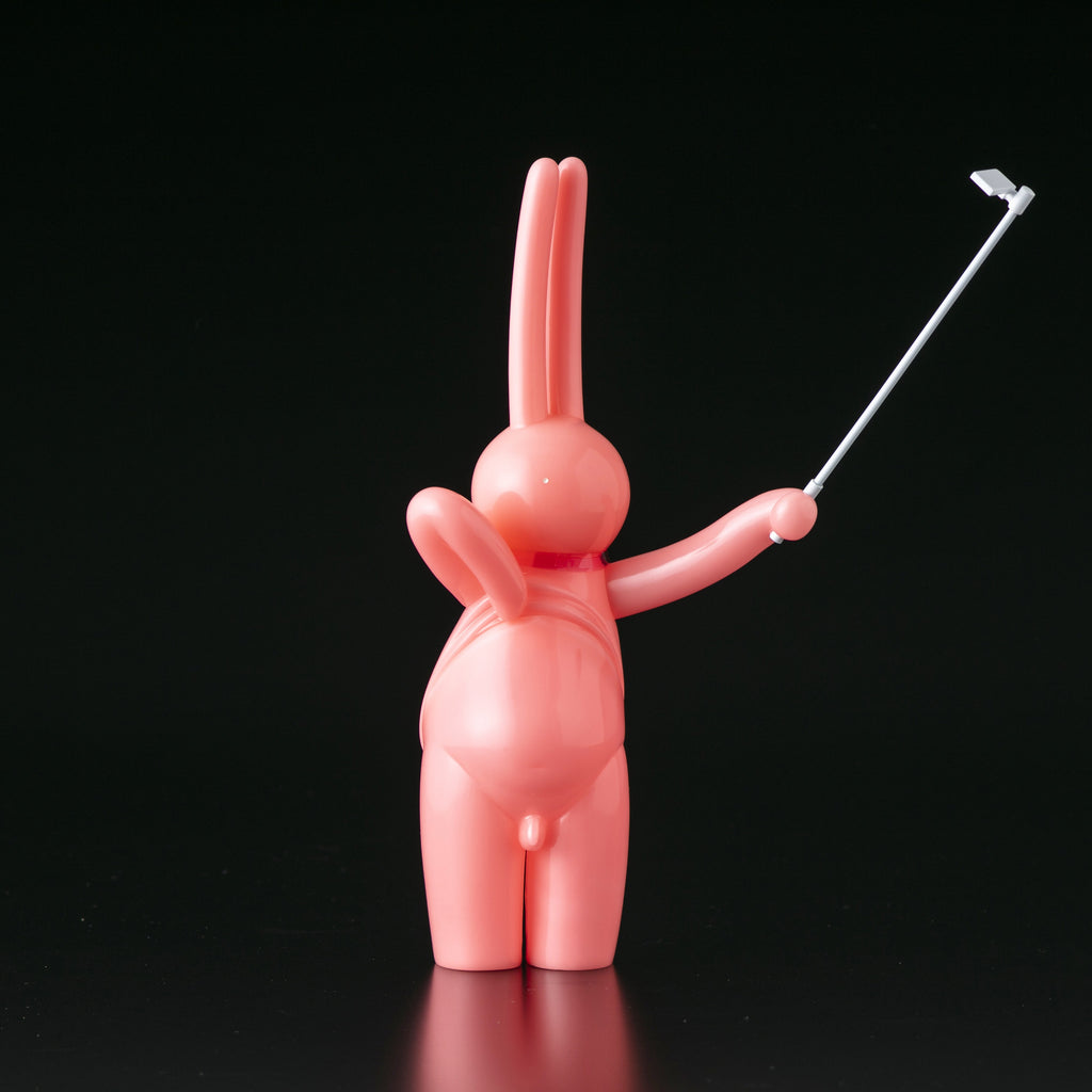 The Daily Flasher by mr. clement (Tomenosuke Shoten) is a pink, glossy sofubi bunny figurine holding a selfie stick upright against a plain black background.