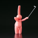 The Daily Flasher by mr. clement (Tomenosuke Shoten) is a pink, glossy sofubi bunny figurine holding a selfie stick upright against a plain black background.