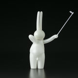 The Daily Flasher by mr. clement for Tomenosuke Shoten is a glossy white bunny figurine, standing upright with a raised right arm holding a selfie stick against a black background.
