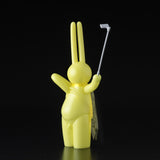 The Daily Flasher by mr. clement for Tomenosuke Shoten is a yellow, long-eared bunny vinyl figure with a selfie stick and translucent cape that glows in the dark—adding playful charm to any collection.
