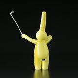 A yellow, bunny-shaped The Daily Flasher by mr. clement from Tomenosuke Shoten stands upright, holding a selfie stick with its left arm against a plain black background—a charming glow-in-the-dark vinyl sofubi figure.