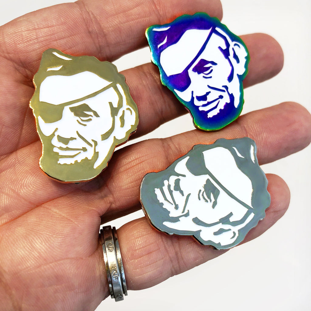 A hand displays three Rotofugi Pirate Abe Enamel Pins—Abraham Lincoln’s face with an eyepatch, in gold, silver, and iridescent finishes. Unique mascot pins from top designer toy brand Rotofugi.