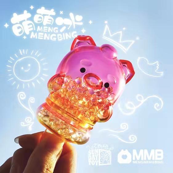 A hand holds a transparent pink Pig Popsicle Mini-Figure from Sun Day Toy’s Meng Meng Bing line, filled with beads; white sun, cloud, and crown doodles decorate the background, with Sun Day Toy and MMB logos visible.
