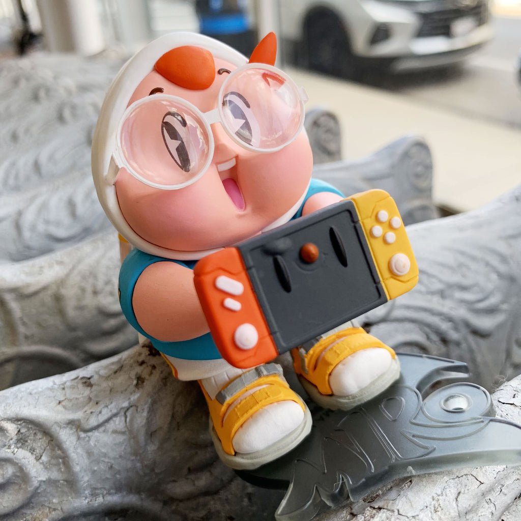 A Miro & Wujo Blind Box figure by LAM Toys, wearing glasses and a chicken hat, holds a Switch-style game console while seated on a decorative silver surface.