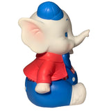 Side view of the Carnival Stripes Baby Elephant plush by Bitter Squeaks, wearing a blue cap, red jacket, blue pants, and white shoes, sitting and pointing its right arm forward.