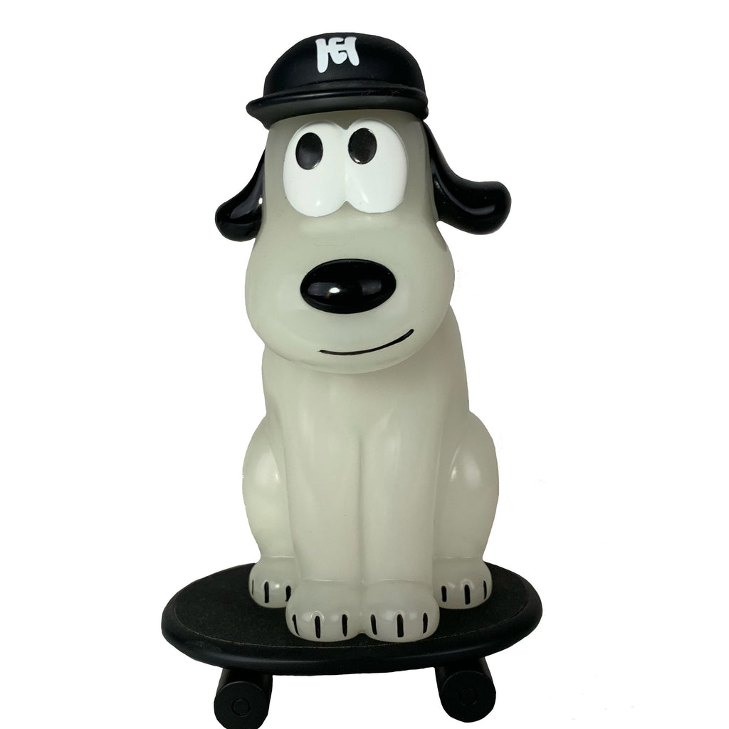 The One-Chu SK8 - Glow by How2Work is a glow-in-the-dark cartoon dog figurine in a black cap, sitting on a skateboard with four wheels against a white background.