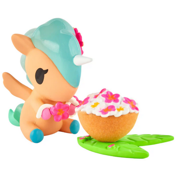 A tokidoki Hawaiian Ohana Blind Box figurine with a blue mane and pink flower sits by a cupcake with white icing and pink flowers on green leaves—a charming addition to any tropical collectibles display.