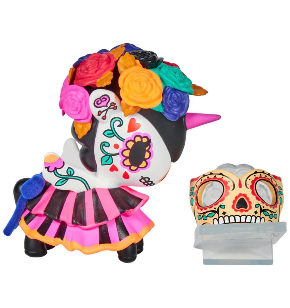A tokidoki Unicorno After Dark Masquerade Blind Box figure, featuring floral and heart patterns, a flower crown, pink horn, and striped skirt, stands next to a decorative Day of the Dead skull mask.
