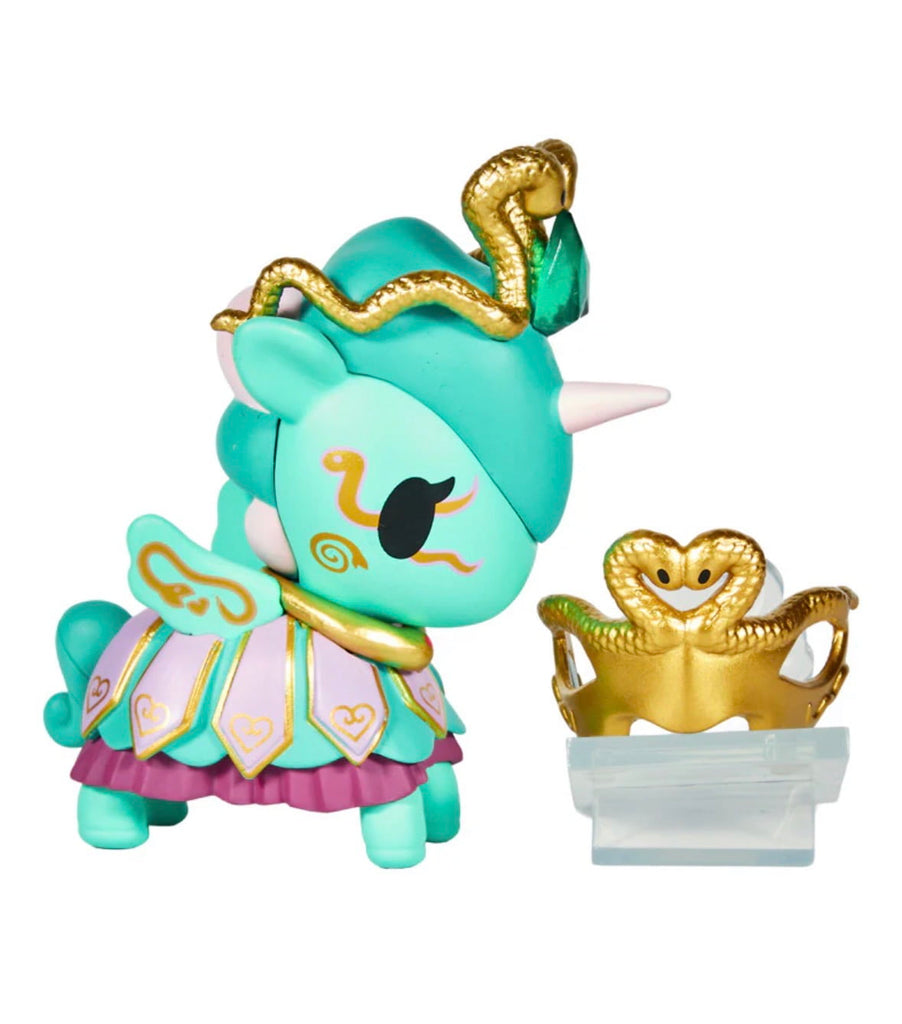 A tokidoki Unicorno After Dark Masquerade Blind Box figurine with teal, gold, and purple accents stands beside a gold crown with a snake motif, both displayed as collectible toys on a clear stand.