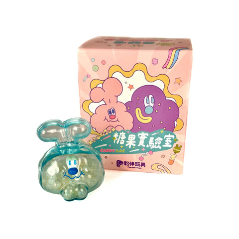 A small blue plastic toy figure with a cartoon face sits before a pastel pink KINGJUN Candy Lab Blind Box by Partner Toys, featuring colorful fruit characters and Chinese text—perfect for blind box figure collectors.