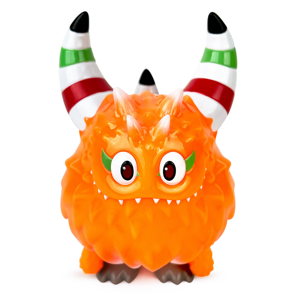 Rangeas Rex Jr. — Lollipop Orange by The Little Hut is a small orange toy monster with large striped horns, green eyelids, two small head horns, wide eyes, pointed teeth, and gray feet on a white background.