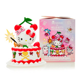 A Hello Kitty figurine with cherries on its head sits in a decorative cake beside a pink tokidoki Tokidoki x Hello Kitty 50th Birthday Blind Box.