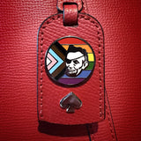 The Rotofugi Abe Progress Pride Hard Enamel Pin, by Rotofugi, features a stylized face with Progress Pride flag colors on a round pin attached to a red leather tag with a spade symbol.