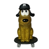 The How2Work One Chu Sk8- Gold features a gold glitter dog figure wearing a black cap, sitting on a black skateboard with gray wheels, facing forward.