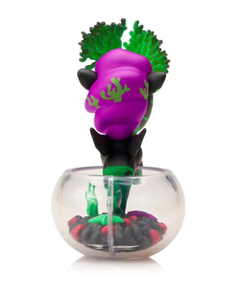 The tokidoki Neon Reef Mermicorno - Deep Star Coral Special Edition features a figurine with purple and green hair in a clear fishbowl, standing on black, red, and purple pebbles, plus glow-in-the-dark accents.