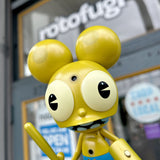 A gold Dalek Space Monkey vinyl figure by UVD Toys, a Rotofugi exclusive, stands in front of a glass door with 