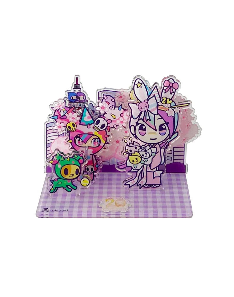 The tokidoki 20th Anniversary Acrylic Standee Blind Box features vibrant cartoon characters with party hats and pastel accents on a purple checkered base, set against a cityscape and cherry blossoms, celebrating 20 years of tokidoki.