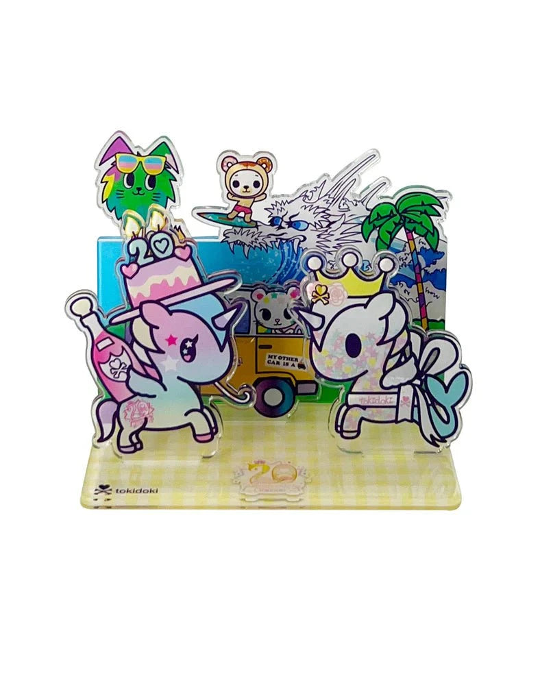 Two cartoon unicorns with party items stand before a colorful Tokidoki 20th Anniversary Acrylic Standee Blind Box backdrop from tokidoki, featuring a cat with sunglasses, a bear, palm trees, a food truck, and a mountain.