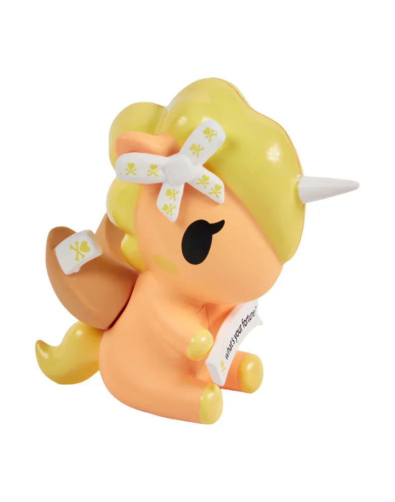 This tokidoki Fortune Cookie Unicorno Blind Box includes a small vinyl unicorn figurine with blonde hair, white horn, brown wings, and a white bow with yellow skulls, sitting while holding a white banner.