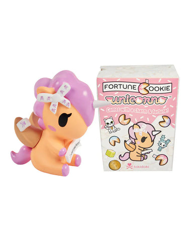 A toy unicorn with a fortune cookie theme is displayed beside the pink "Fortune Cookie Unicorno Blind Box" by tokidoki, decorated with cute illustrations and text. Open it to reveal a surprise unicorn figure hidden inside!.