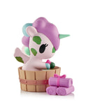A pastel Spa Day Unicorno Blind Box figurine by tokidoki, featuring a green horn, relaxes in a wooden tub beside three purple towels with a pink ribbon, showcasing kawaii wellness on a white background.