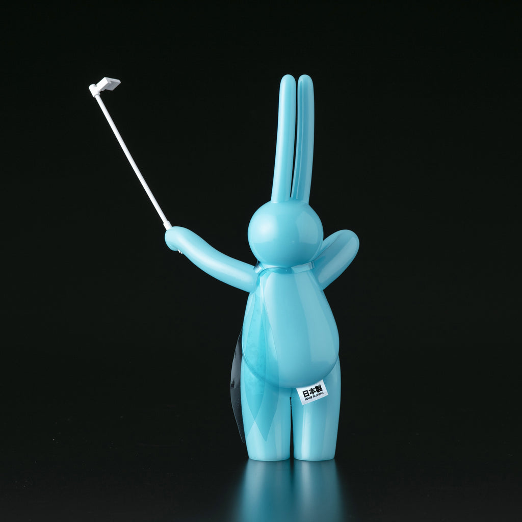 The Daily Flasher by mr. clement, from Tomenosuke Shoten, is a blue cartoon-like rabbit figure standing on two legs and holding a selfie stick in its right hand against a plain black background.