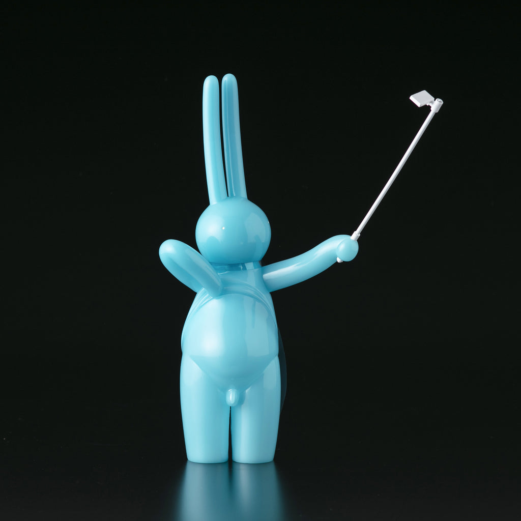 The Daily Flasher by mr. clement from Tomenosuke Shoten is a glossy blue sofubi bunny figure standing upright, holding a selfie stick with a phone attached, set against a plain black background.