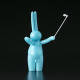 The Daily Flasher by mr. clement from Tomenosuke Shoten is a glossy blue sofubi bunny figure standing upright, holding a selfie stick with a phone attached, set against a plain black background.