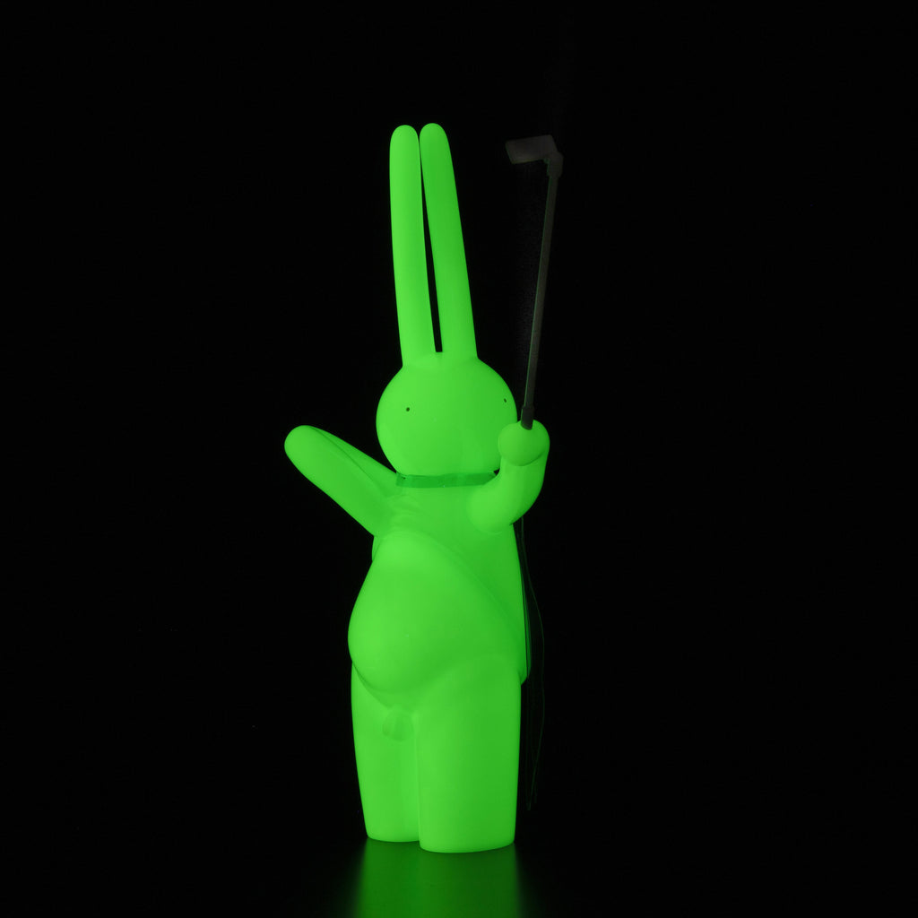 The Daily Flasher by mr. clement, from Tomenosuke Shoten, is a glowing green rabbit vinyl toy that stands upright in the dark, holding a thin stick and capturing the whimsical spirit of Tomenosuke Shoten collectibles.